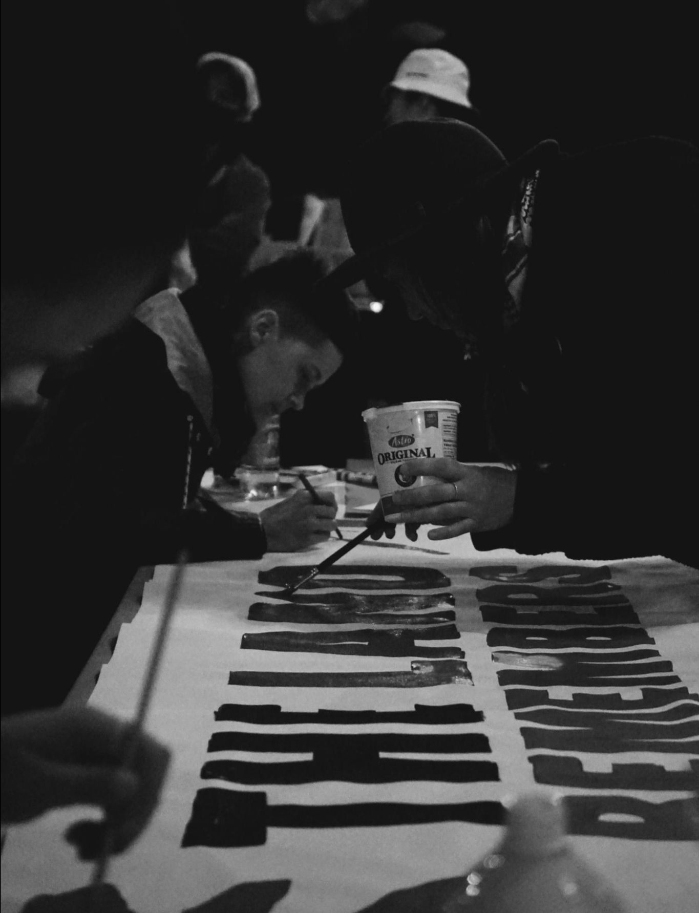 A black and white image of three people painting block letters 'the land remembers' on a banner