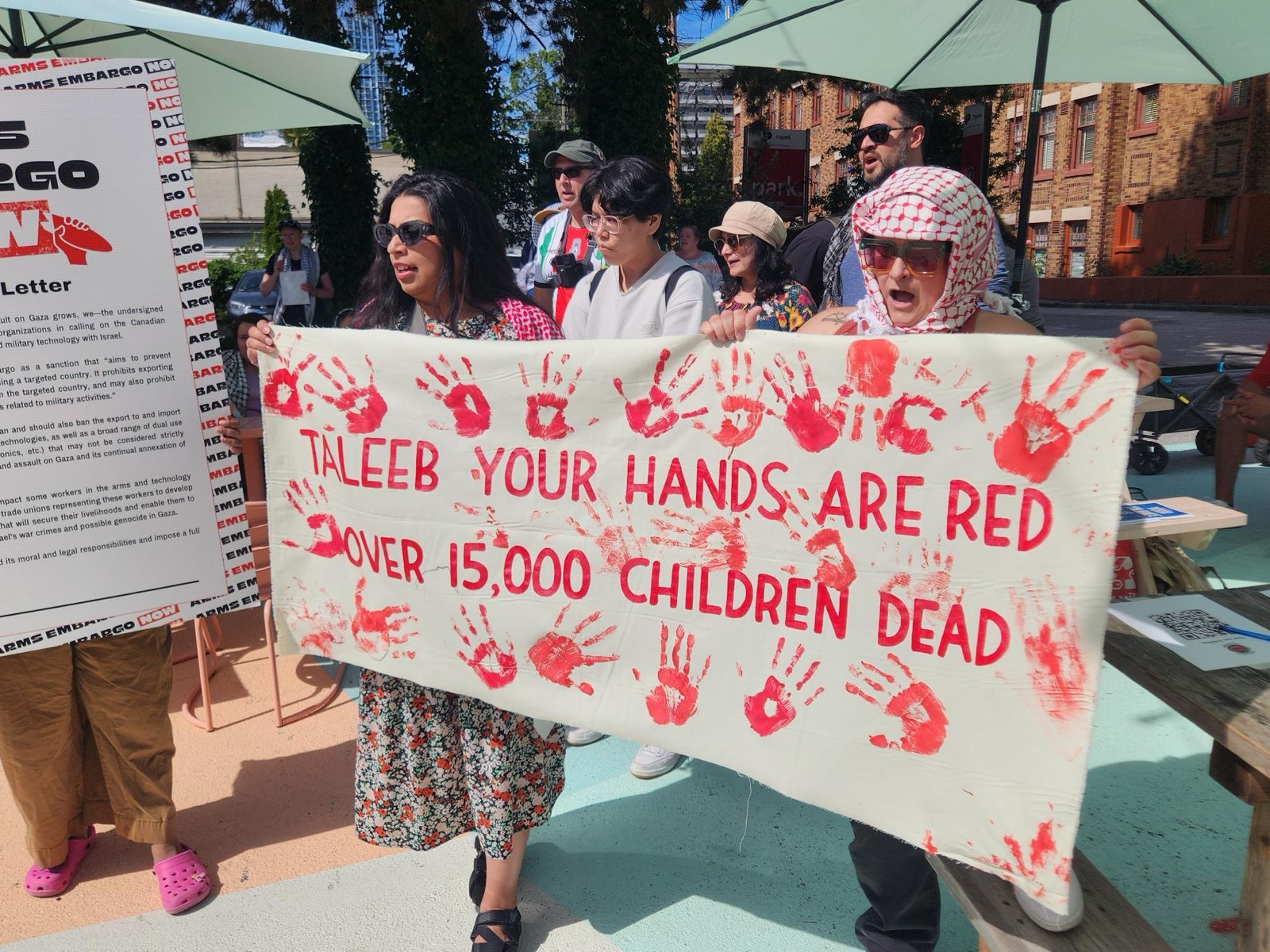 a group of people holding a white banner with red hand prints on it that says Taleeb your hands are read, over 15,000 children dead