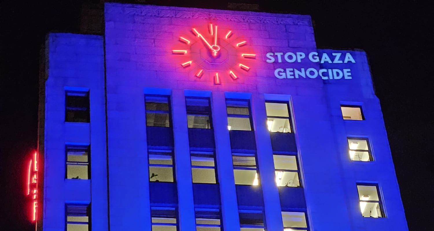 A photo of City Hall lit up in blue with a light projection on the top right that reads stop gaza genocide