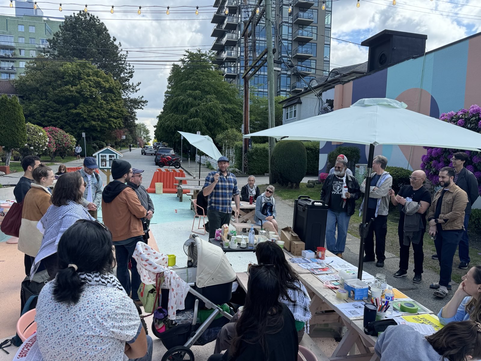 A large group of people standing around picnic tables laid out with coffee and posters